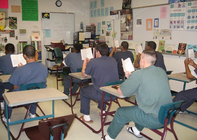 Young people in blue and green clothes sit at desks in a classroom setting, holding books, facing away from the camera.