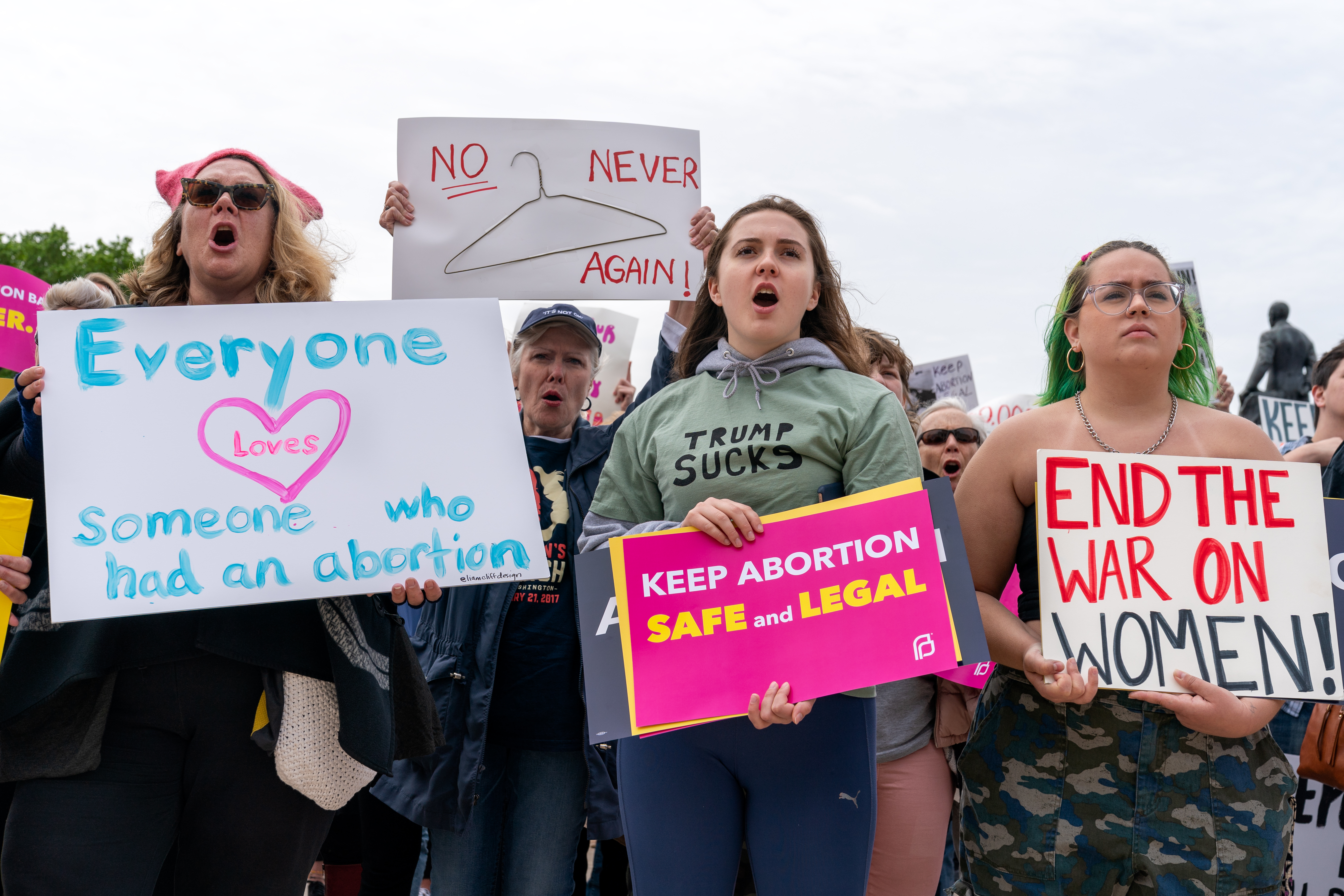 Four women stand at the front of a group of protestors, holding signs that read: "Everyone loves someone who had an abortion;" "No Never Again" with a coat hanger; "Keep Abortion Safe and Legal;" and "End the War on Women!"