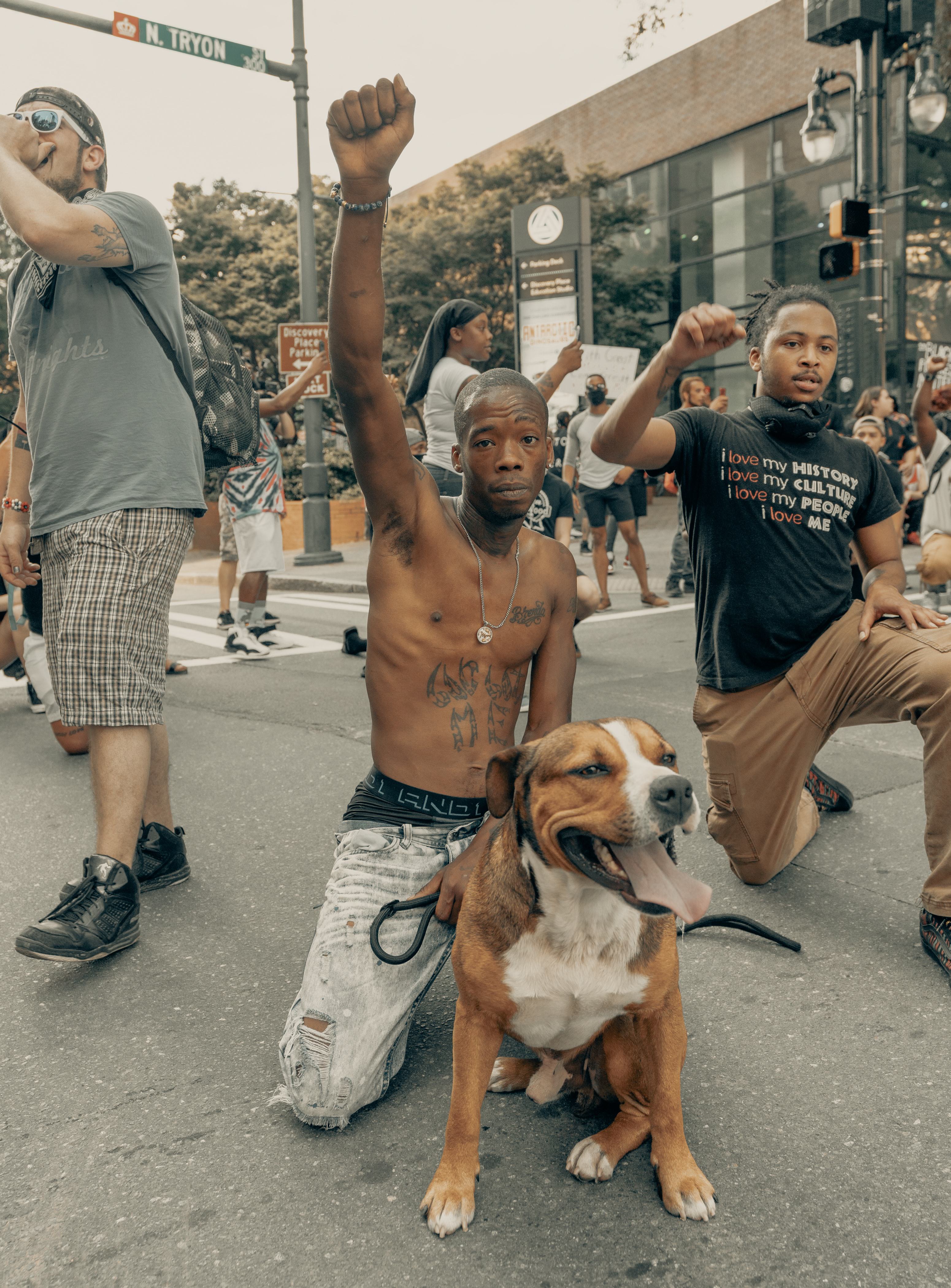 Two black protestors kneel with raised fists in an intersection; one has a dog.
