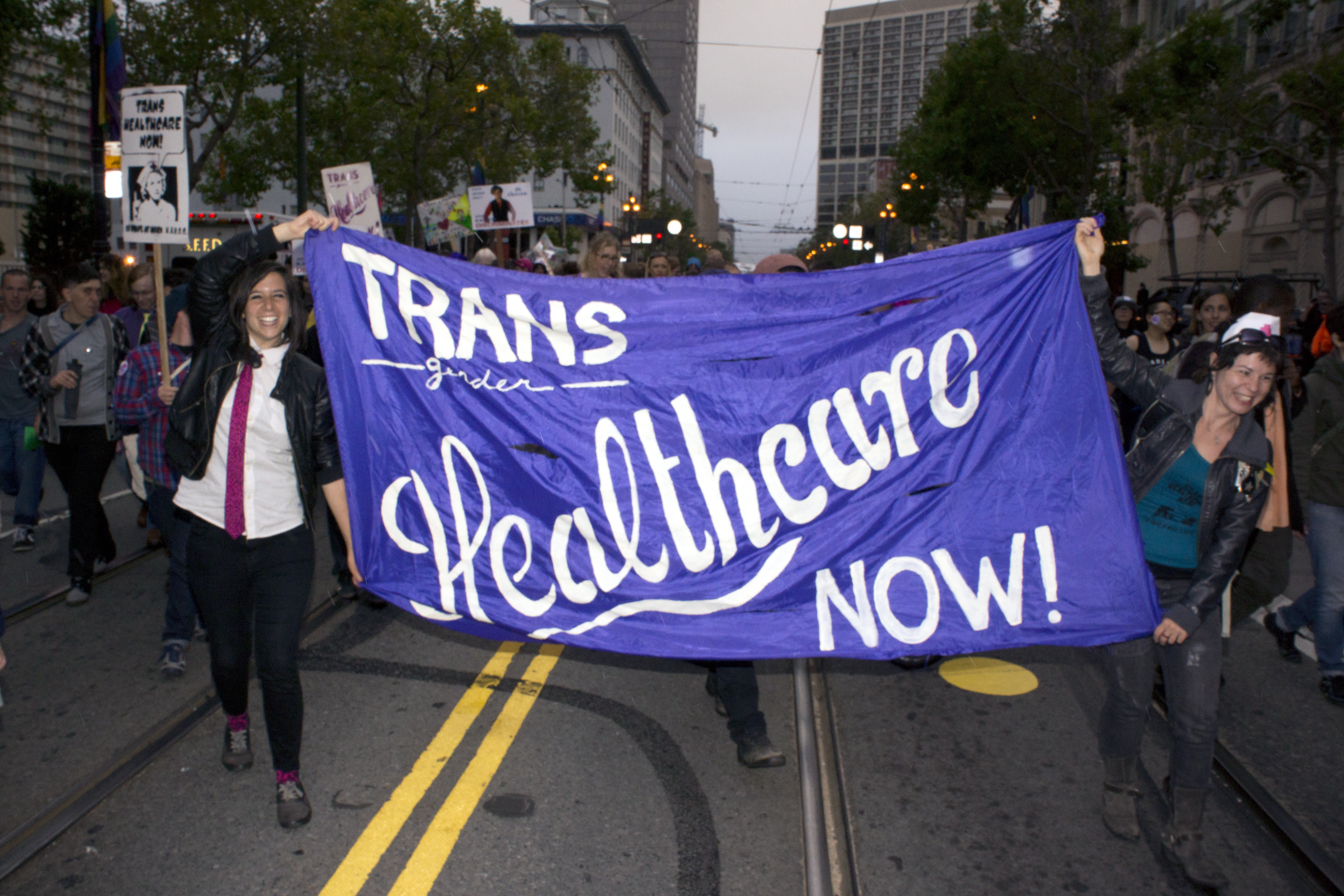Two people walk in front of a group, down a street, holding a banner reading "Transgender Healthcare Now."