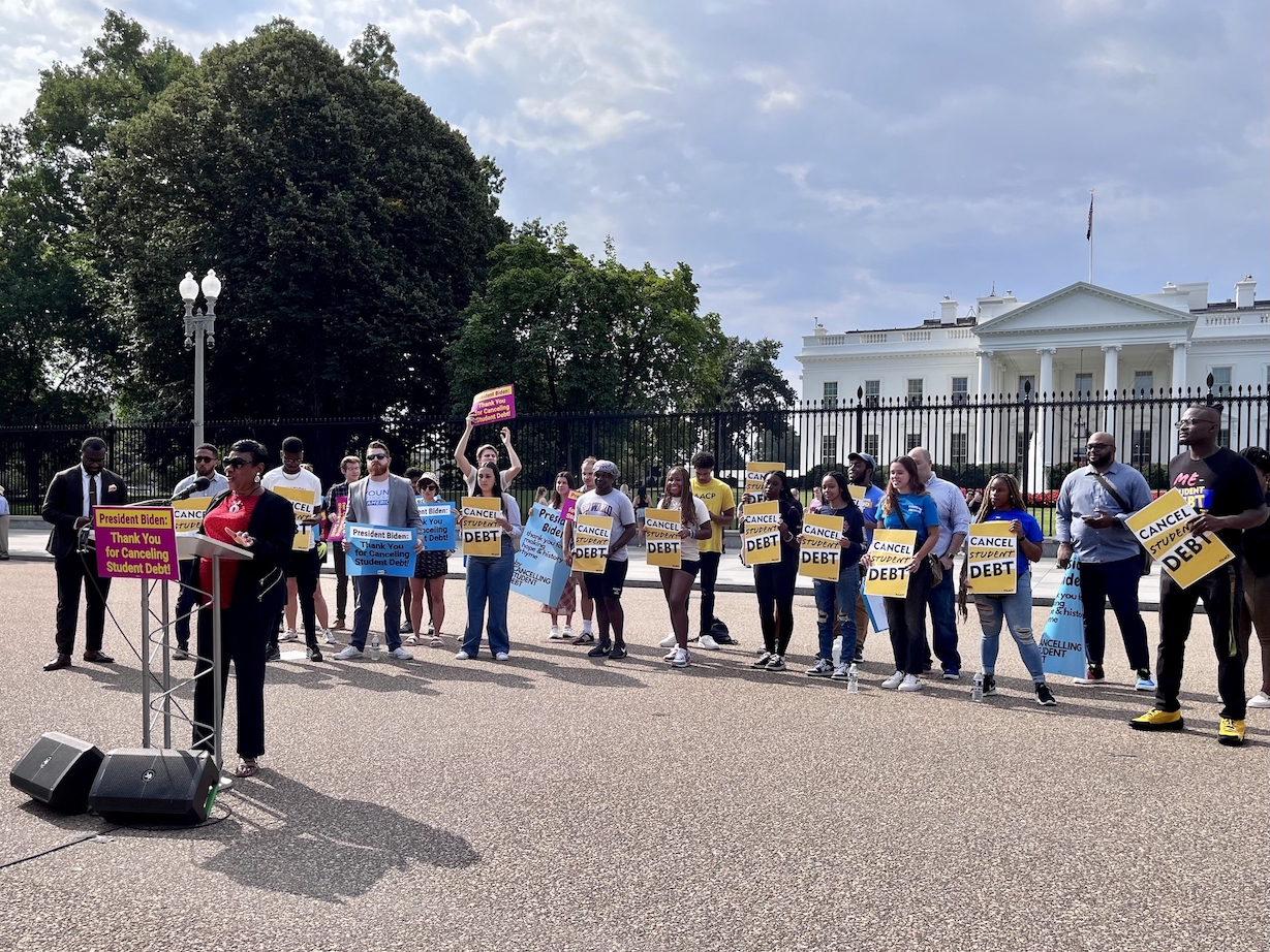 NEA President Becky Pringle stands at podium in front of the White House, with several people behind her holding signs that read: "Cancel Student Debt" and "President Biden Thank You for Canceling Student Debt."