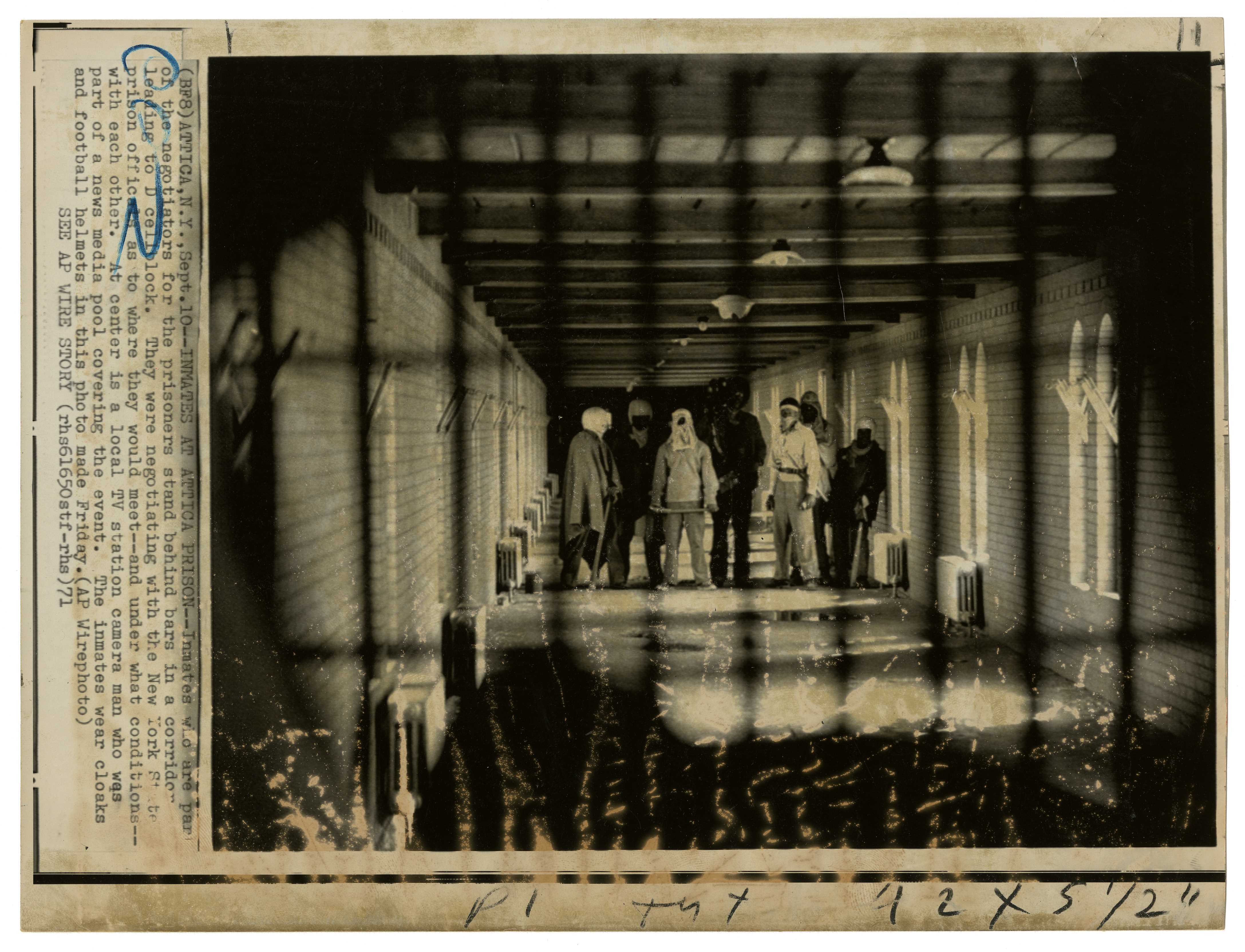 Black and white photo: Attica prisoners participating in the riot stand behind bars in a prison corridor.