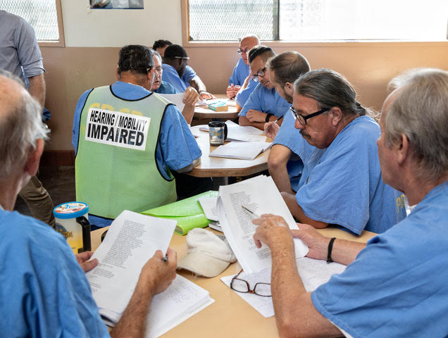 Prisoners in blue sit around tables working; one has a "hearing/mobility impaired" vest