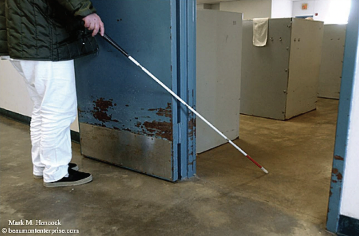 Incarcerated person, visible from mid-torso down, uses a white cane to navigate through a blue prison doorway.
