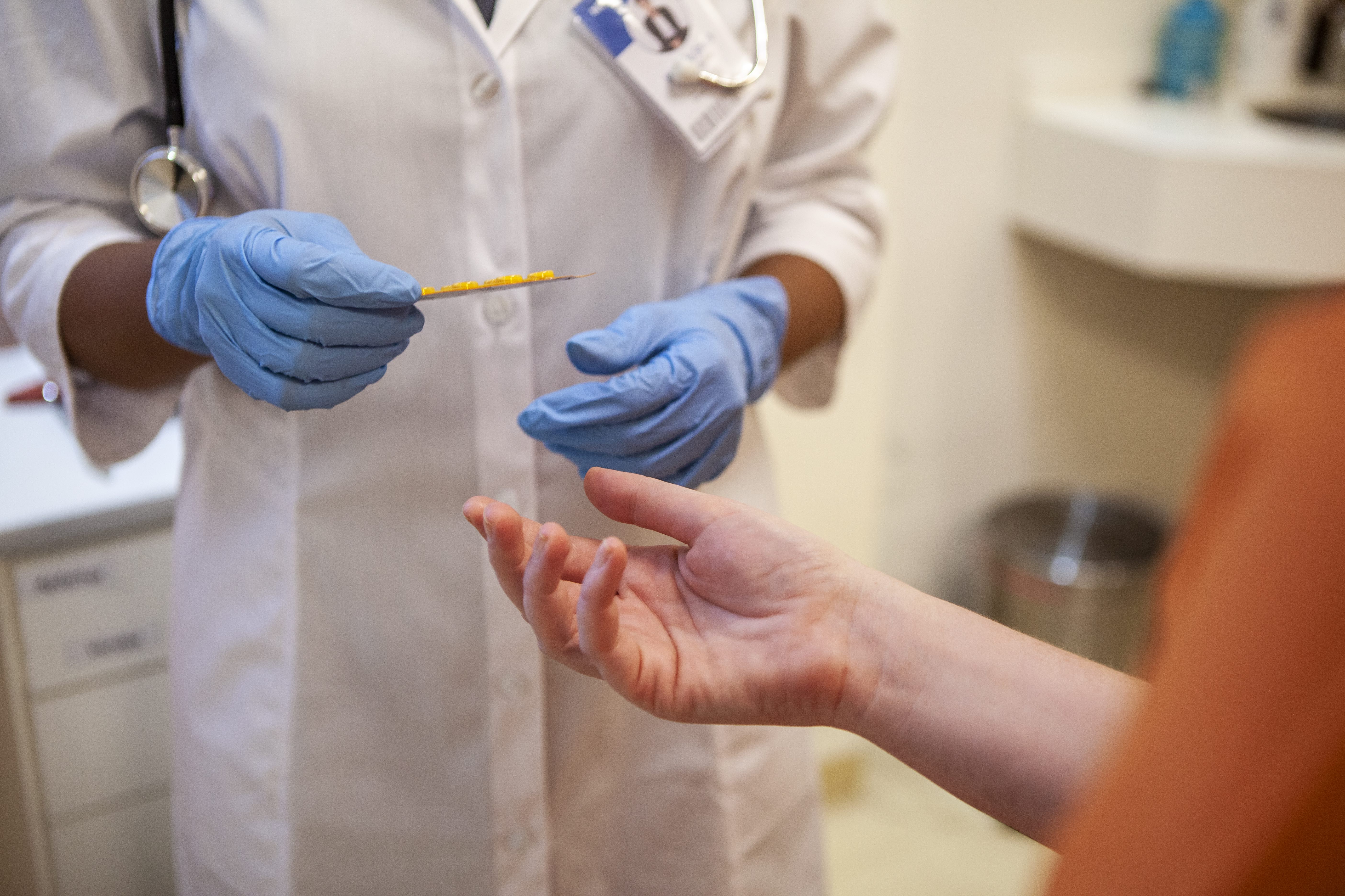 A person in an orange top reaches their hand toward a person in a white coat, with a stethoscope and blue gloves, who is holding a blister pack of medication.